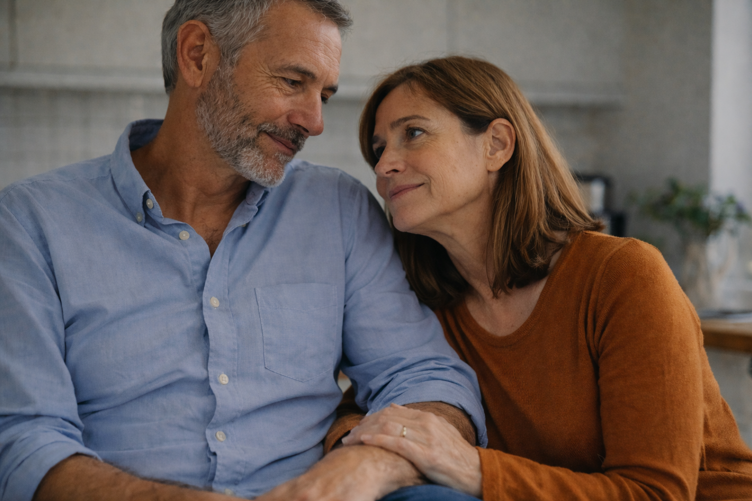 Middle-aged couple sitting closely together in a kitchen, making eye contact with gentle, appreciative smiles, conveying quiet connection and emotional closeness.