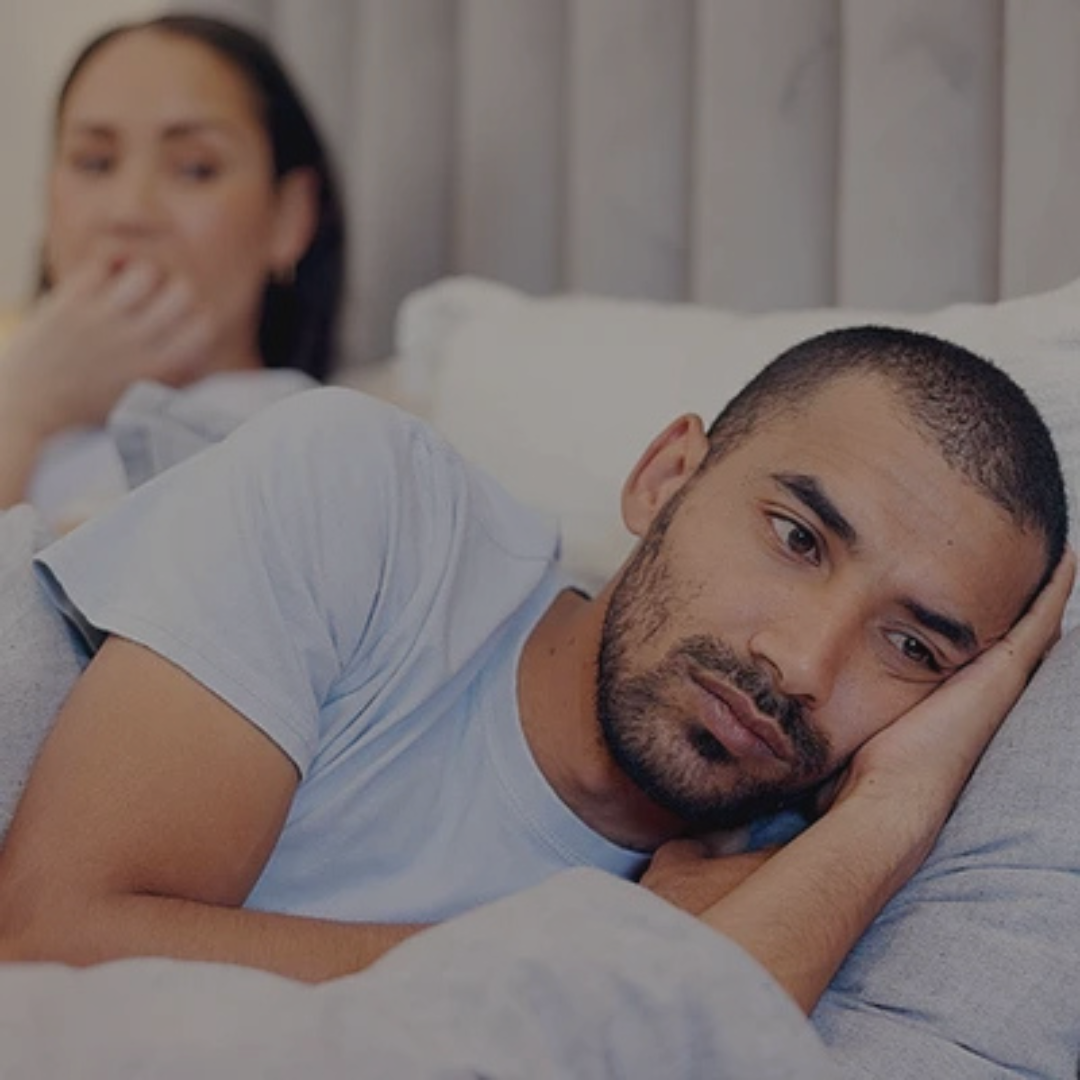 A man lies awake in bed with a pillow, looking pensive, while a woman sits in the background of the bed, blurred. He recently learned of her cheating in an affair.